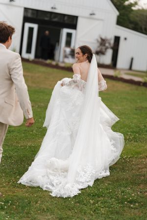 Bride in a lace wedding gown and long veil walks across a green lawn toward a groom in a tan suit near a white barn venue.