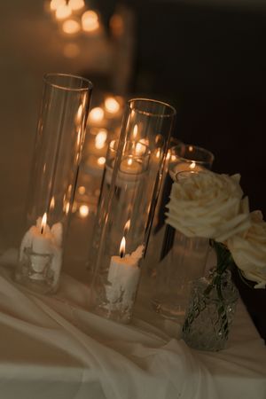 Lit white candles in tall clear glass cylinders beside a cream rose in a vintage vase on a draped table, with warm glowing lights behind.