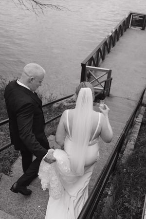 Black and white photo of a bride in a long veil and a groom in a dark suit walking down steps toward a waterfront dock.