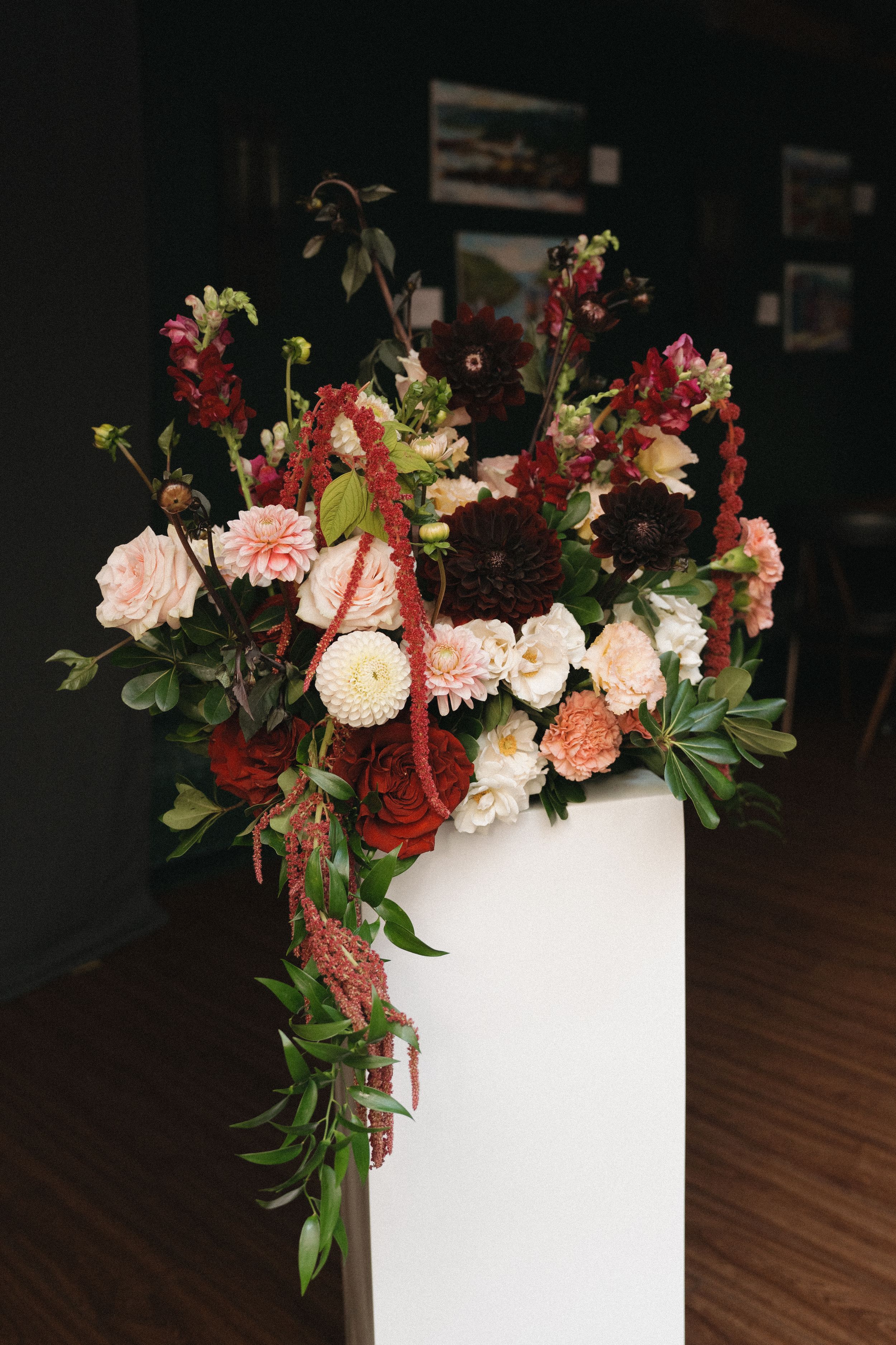 Lush floral arrangement of blush, cream and burgundy dahlias, roses and trailing amaranthus with greenery atop a white pedestal.