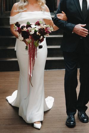 Bride in an off-the-shoulder white gown holds a burgundy and blush bouquet with amaranthus while walking arm in arm with a man in a suit.
