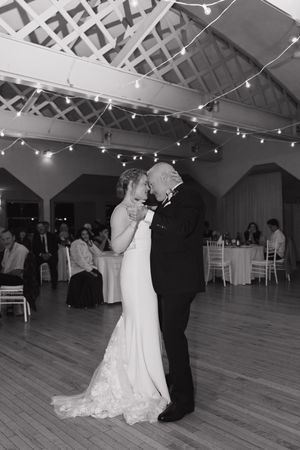 Bride in a lace gown and groom in a dark suit share a first dance under string lights as wedding guests watch from nearby tables.