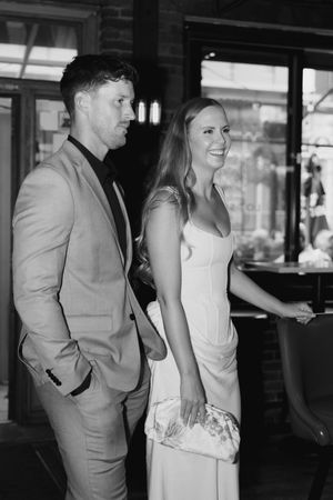 Black and white photo of a smiling couple at an event, the man in a suit and the woman in a long gown holding a floral clutch.