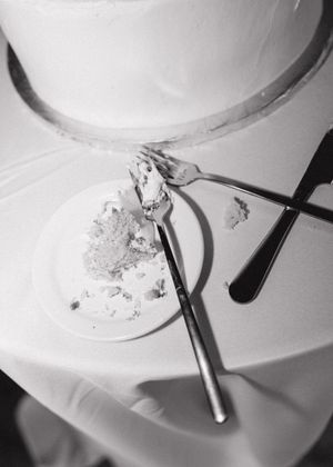 Black and white photo of a wedding cake on a draped table with a partly eaten slice on a plate and forks crossed beside it.