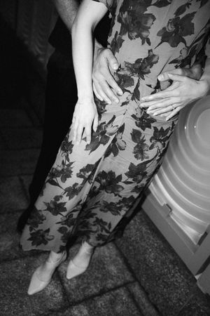 Black and white photo of a woman in a floral jumpsuit and pointed flats, with a partner's hands resting on her waist.