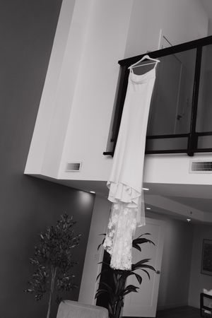 Black and white photo of a wedding dress with a lace train hanging from a balcony railing above potted plants in a modern interior.