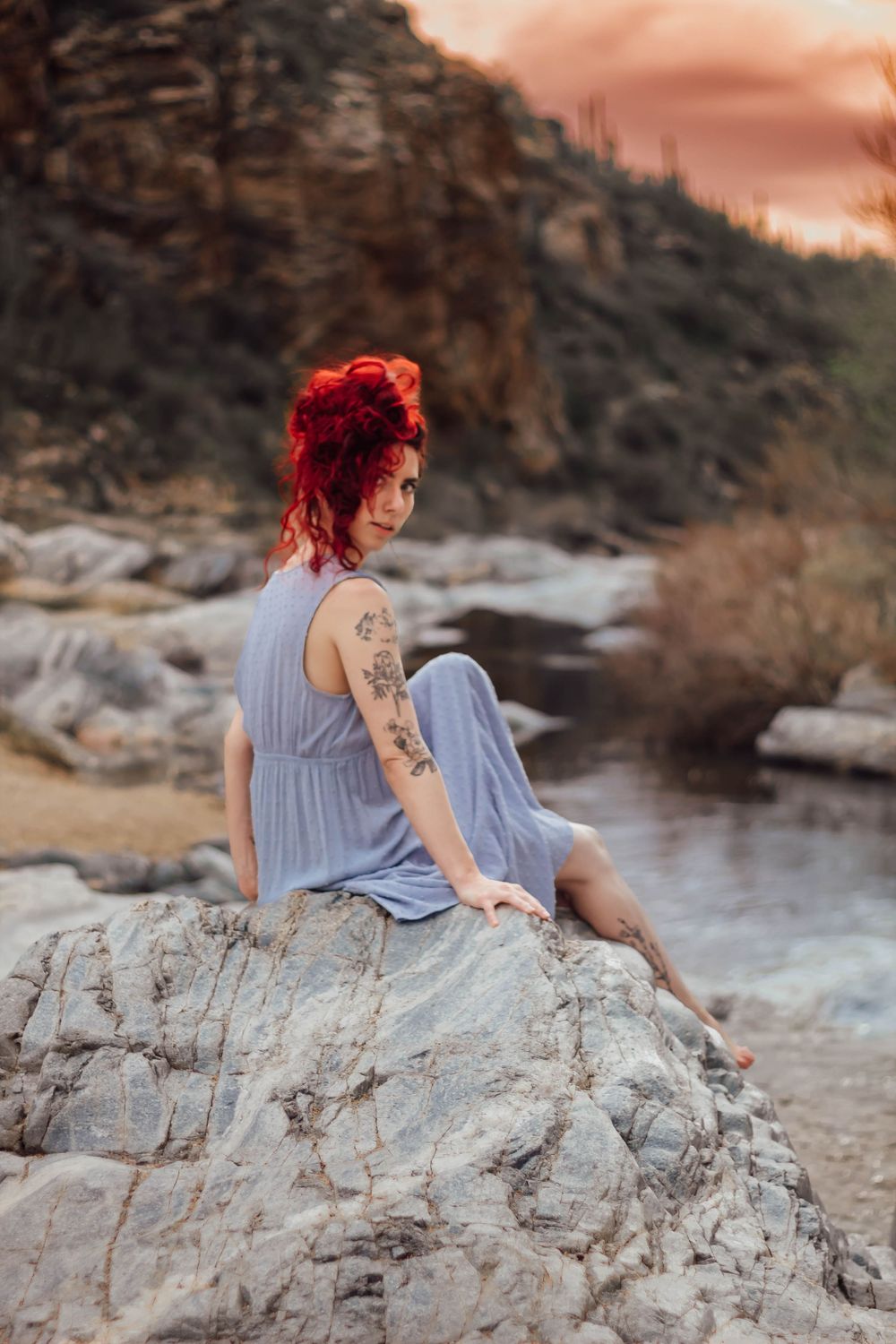 Woman poses for solo portrait session on rocks with a desert sunset in background at Tanque Verde falls in Tucson, Arizona.