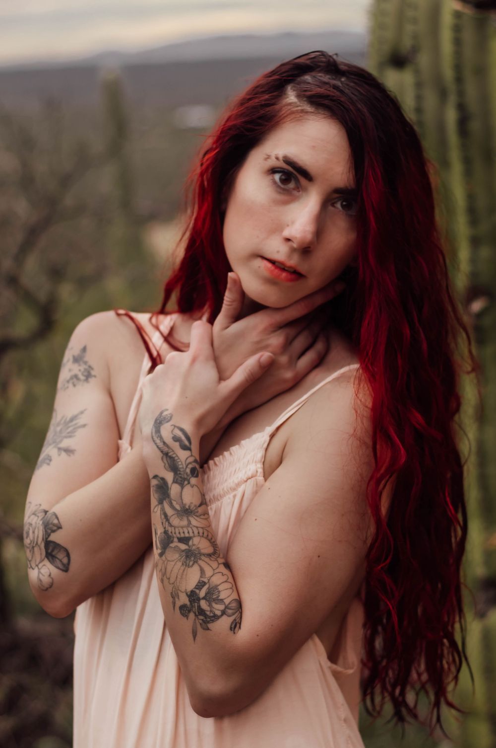 Redhead woman stares at camera while clutching neck, with a cactus in background.