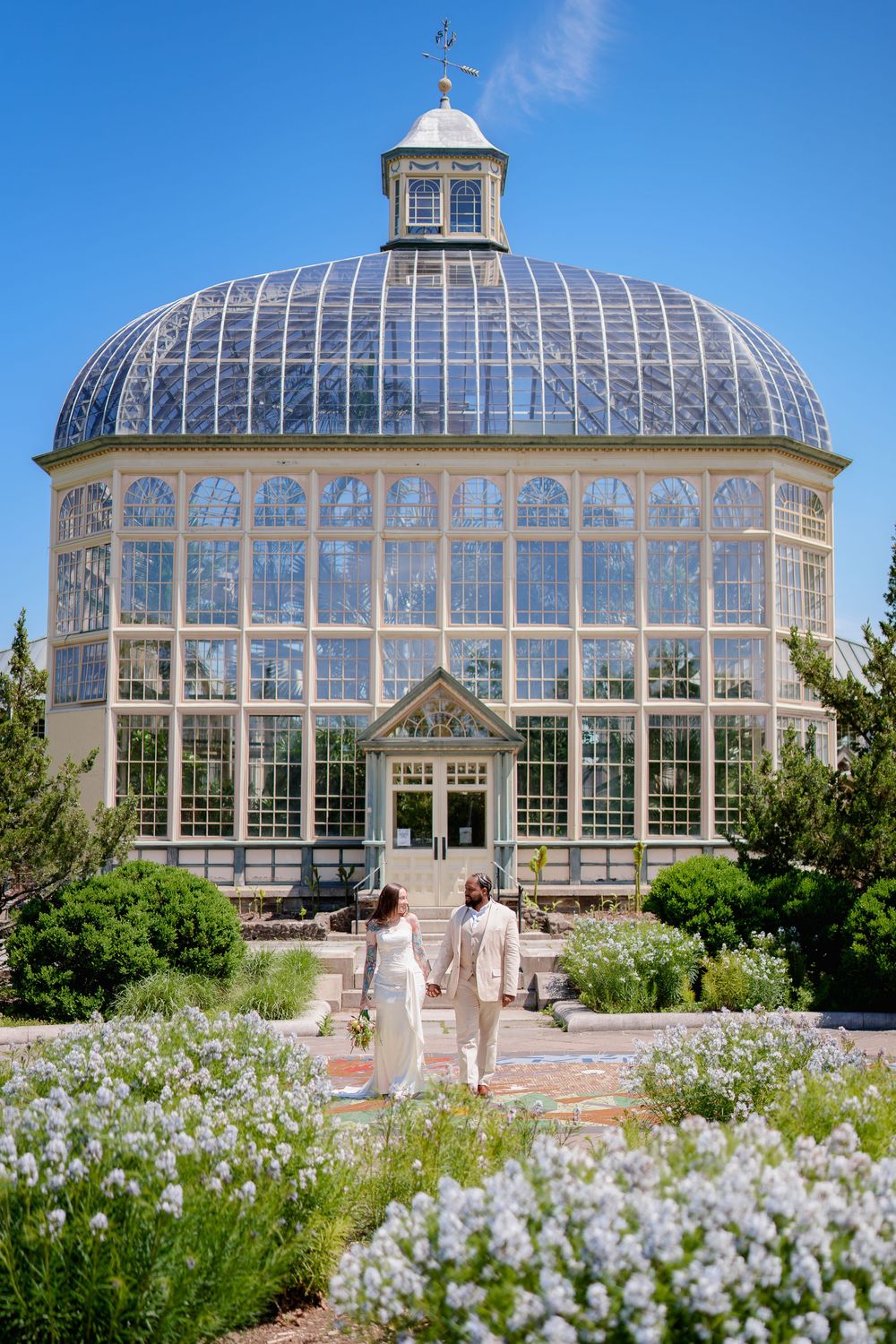 Couple walks hand-in-hand toward a stunning Victorian glass greenhouse conservatory surrounded by blooming gardens under blue sky, in MD