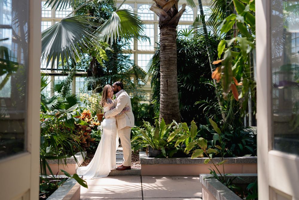 A couple shares a romantic kiss in a lush greenhouse wedding venue surrounded by tropical plants and palm trees, Baltimore, MD