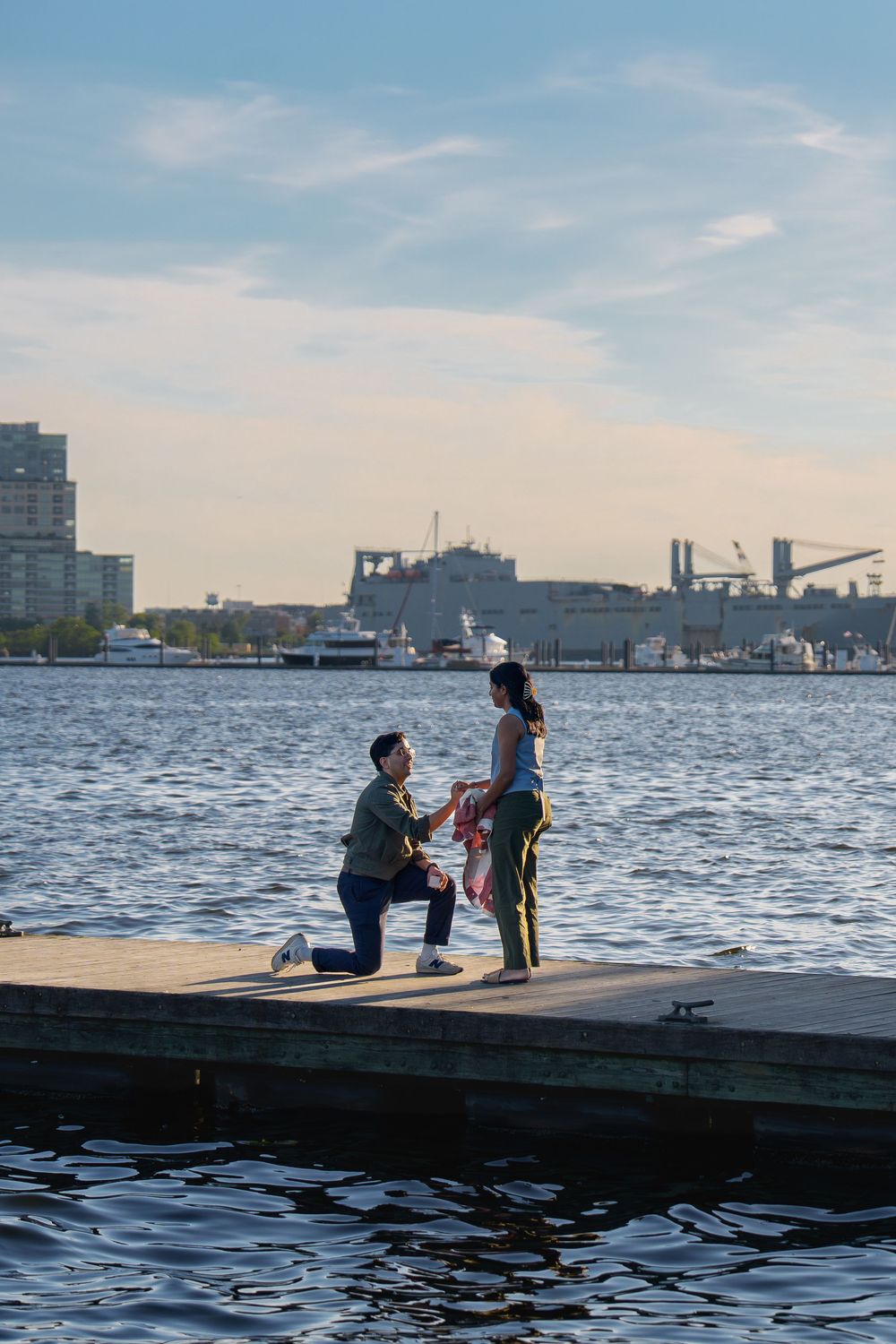 Man proposes to woman on a waterfront dock with city skyline and harbor in the background, in Baltimore, MD