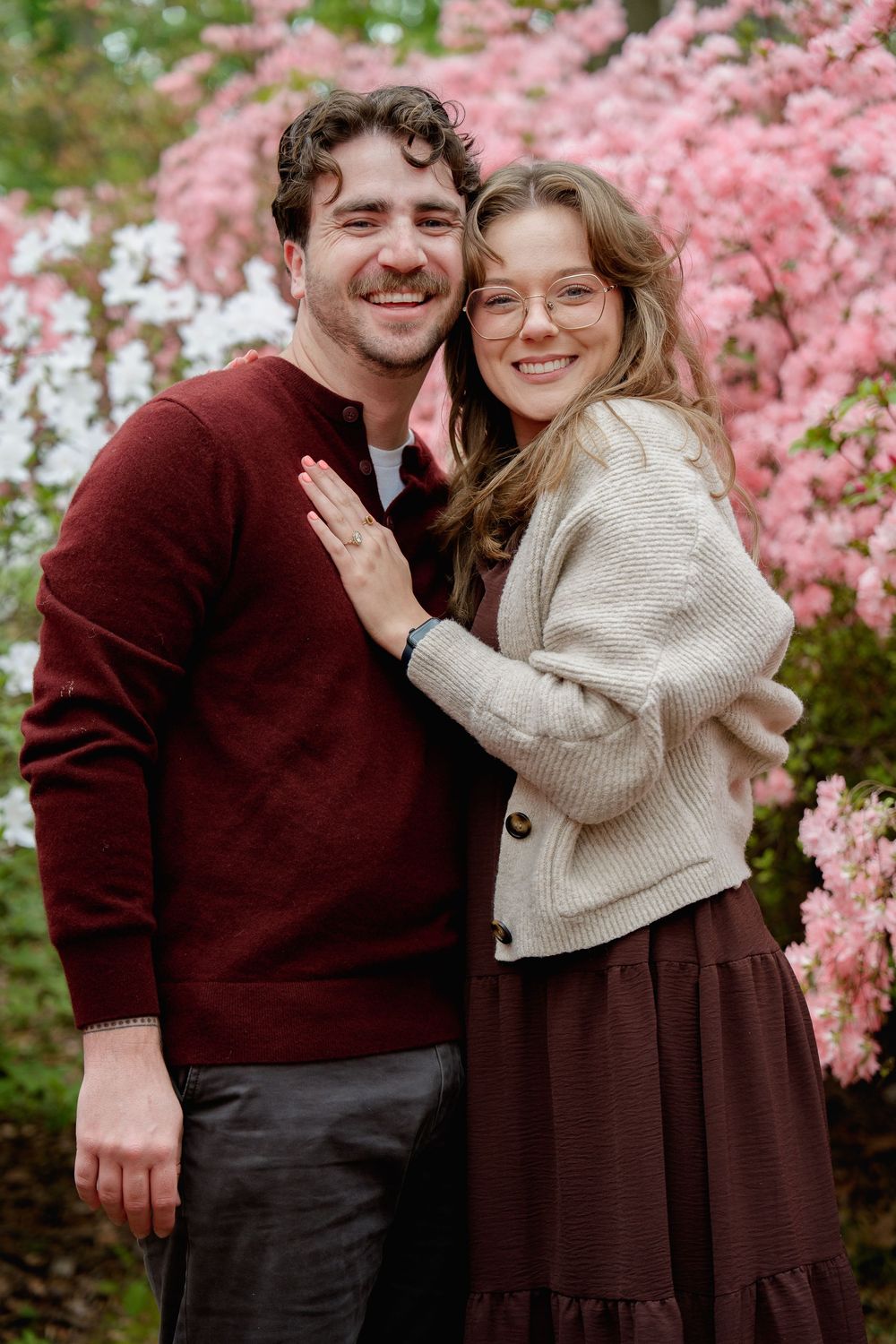Happy couple embracing outdoors near pink flowering trees, woman wearing glasses and showing engagement ring, during proposal session, in MD