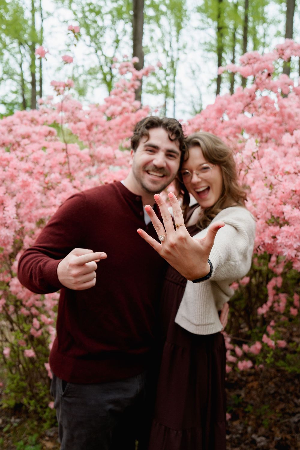 Happy couple showing off engagement ring in front of pink flowering bushes, smiling and pointing at the diamond ring, in Brookeville, MD