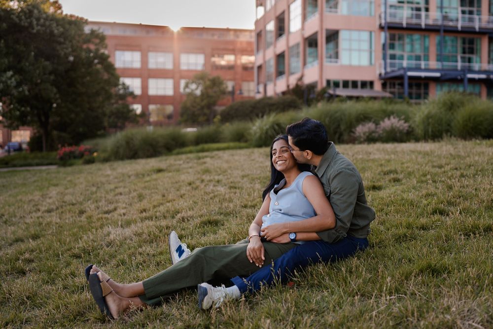 Engage couple sitting together on grass at sunset, woman leaning back against man near urban building backdrop in Baltimore, MD