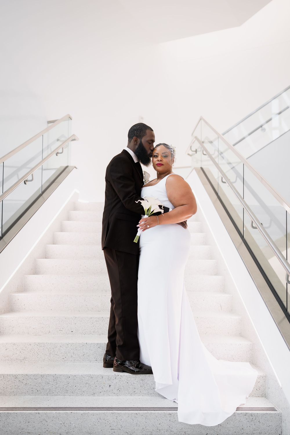 A couple in wedding attire embraces on white marble stairs, bride in white gown holding flowers, in Ellicott City, MD