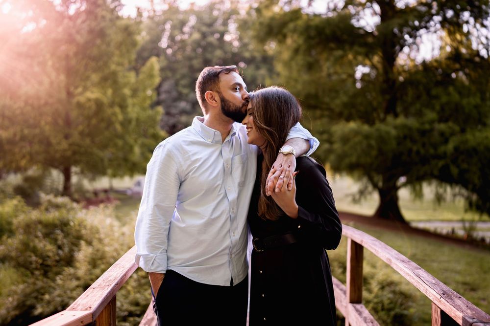 Couple sharing a romantic kiss on a wooden bridge surrounded by lush green trees in a sunlit park, in Wheaton, MD