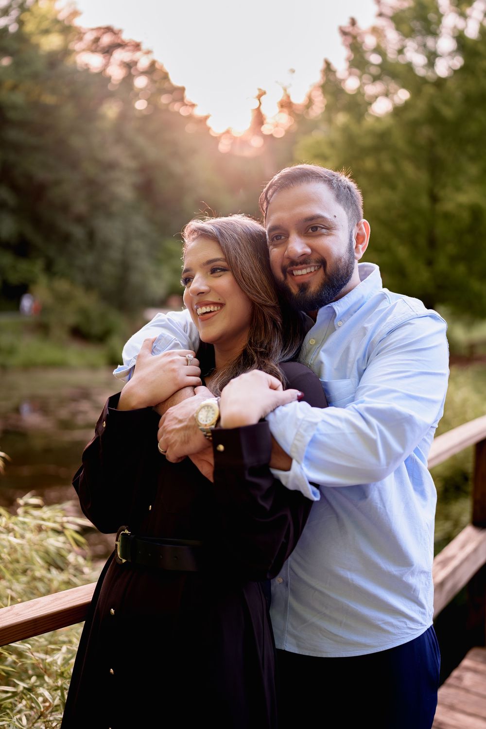 A smiling couple embraces on a wooden bridge in a sunlit park, the man hugging the woman from behind, in Brookside Gardens, Wheaton, MD