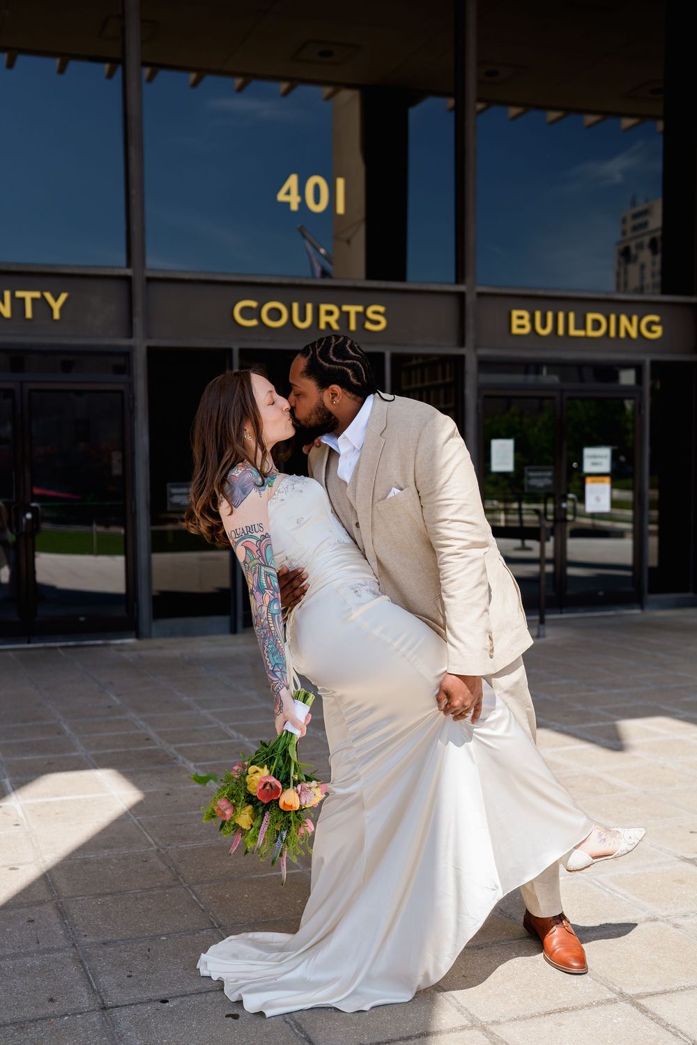 Newlywed couple sharing a romantic dip kiss outside the County Courts Building 401, bride holding colorful bouquet, in Baltimore, MD