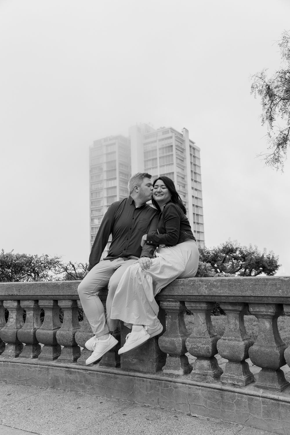 A romantic black and white photo series of a couple sitting together on a stone wall with city buildings in the background.