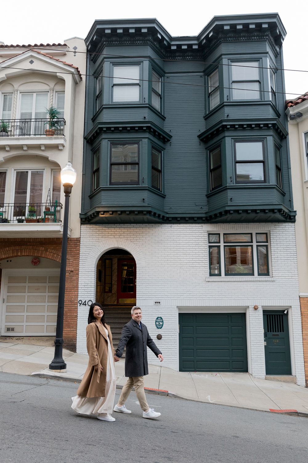 Victorian style houses with bay windows line a steep San Francisco street with people walking past.