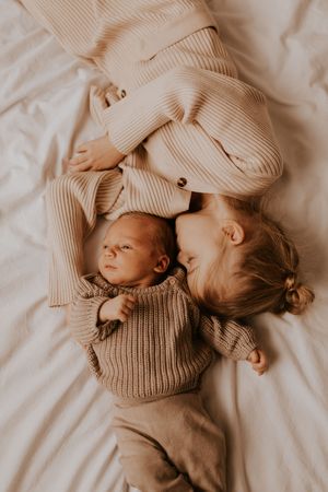 Siblings cuddling together on white bedding wearing cozy beige sweaters.