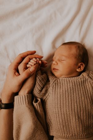 Close-up of tiny baby hand holding adult finger in beige knit sweater.