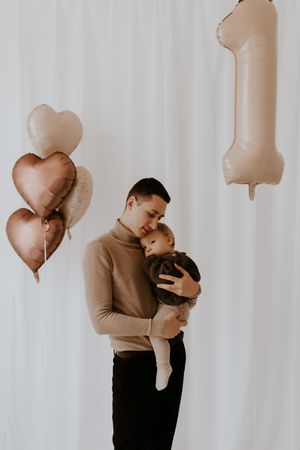 Tender family moment captured in a minimalist setting with beige heart-shaped balloons floating above.