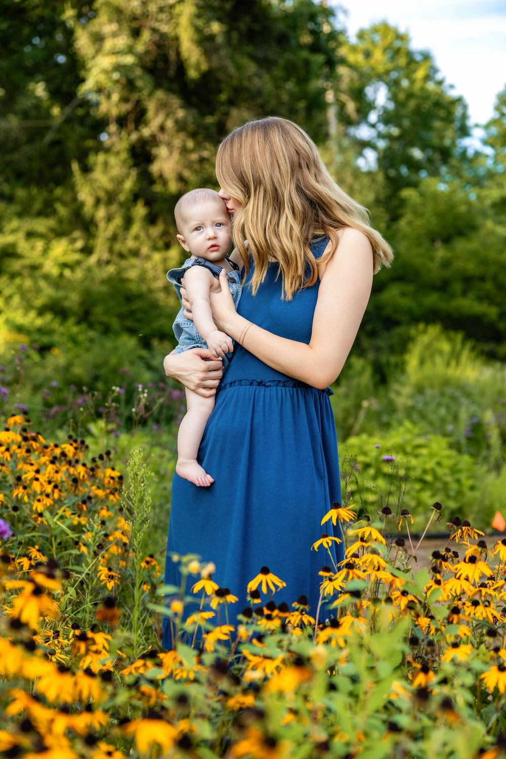 A woman in a blue dress holds a baby among yellow black-eyed Susan flowers in a lush garden.