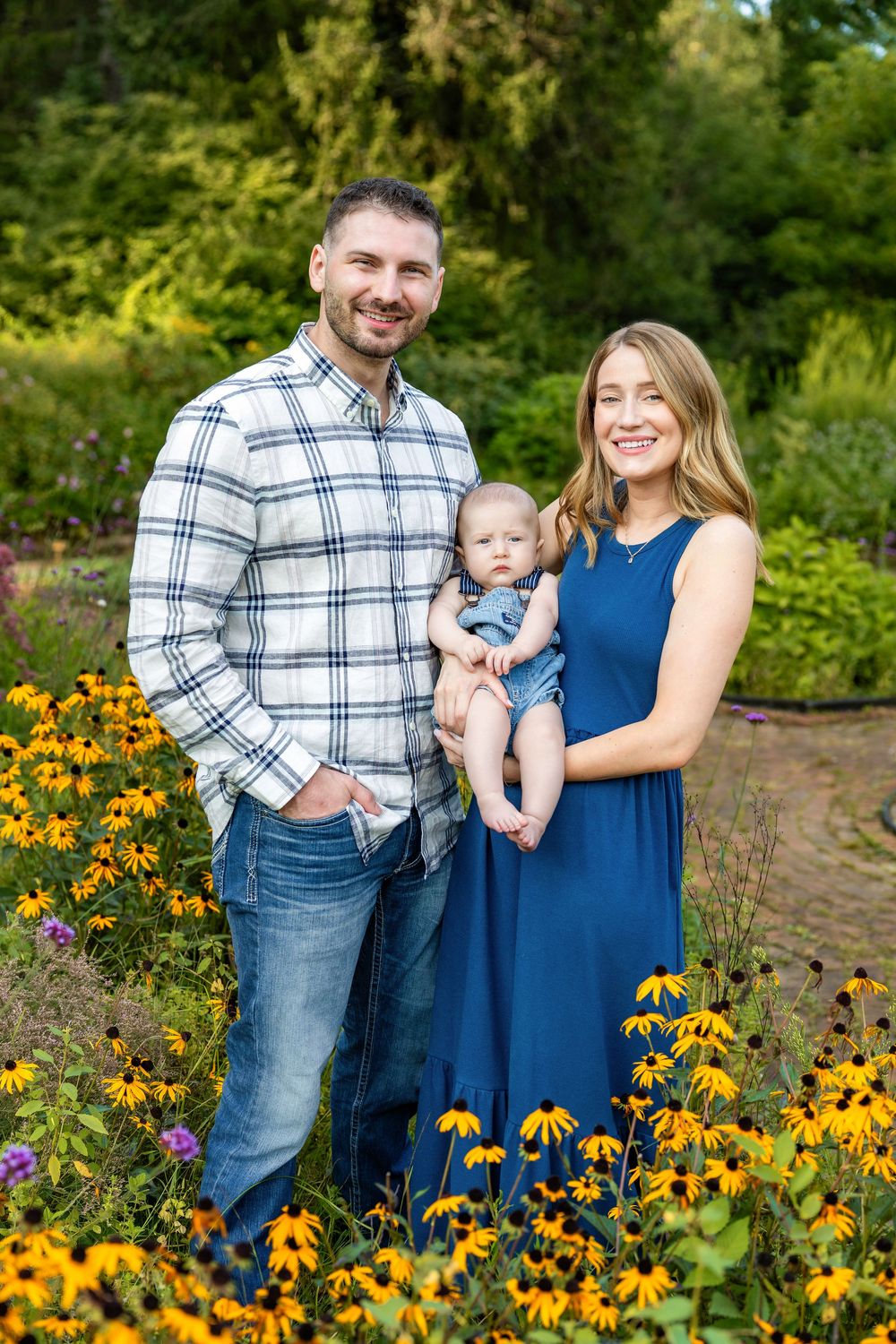 A smiling family with a man in plaid, woman in blue dress, and baby boy posing among yellow black-eyed Susan flowers.
