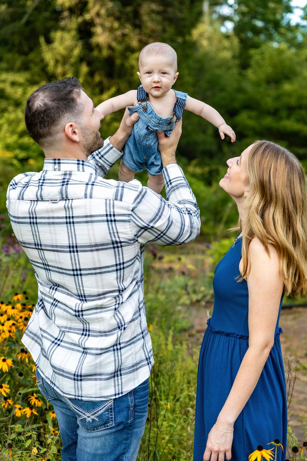 A father lifts his baby in the air while mother looks on, smiling outdoors in a garden setting.