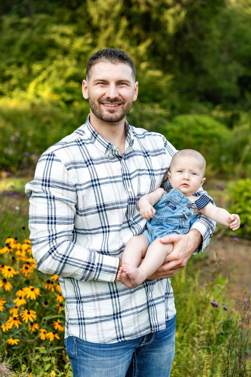 Smiling father in plaid shirt holding baby in denim overalls outdoors with yellow flowers and green trees.