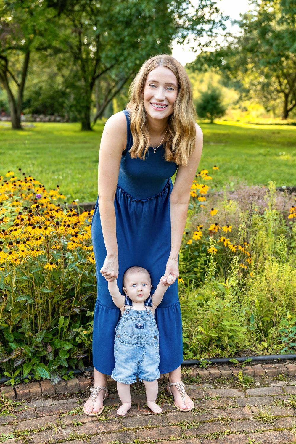 Smiling woman in blue dress holds baby in denim overalls while standing in a lush green park with yellow flowers.