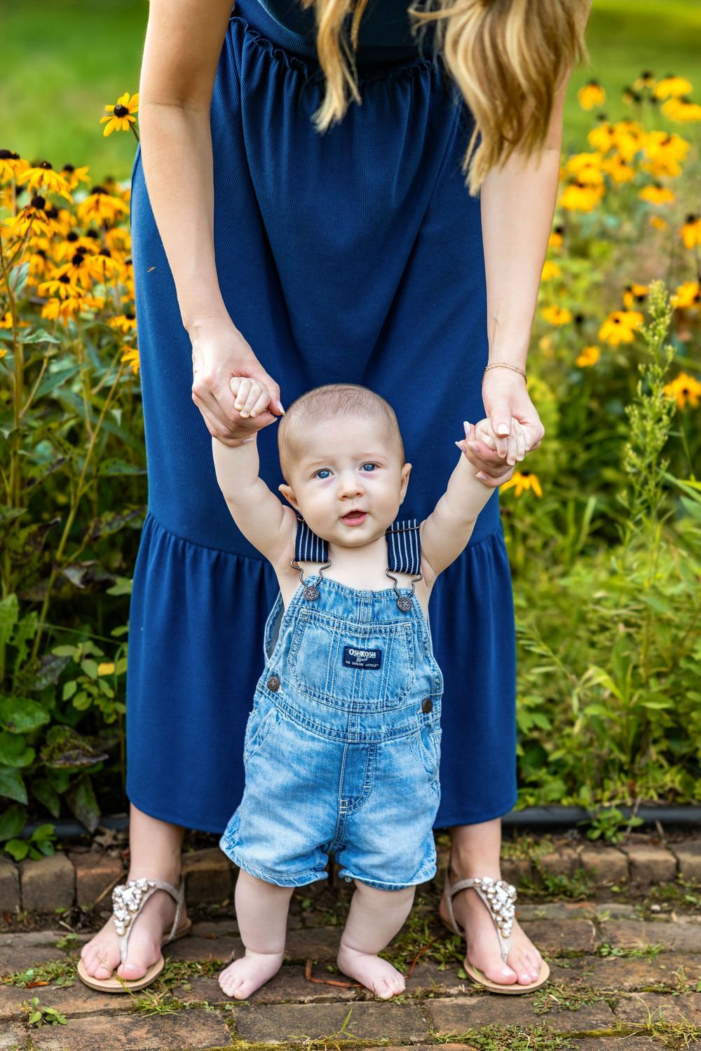 Baby in denim overalls learning to walk with mother in blue dress among yellow flowers in garden.