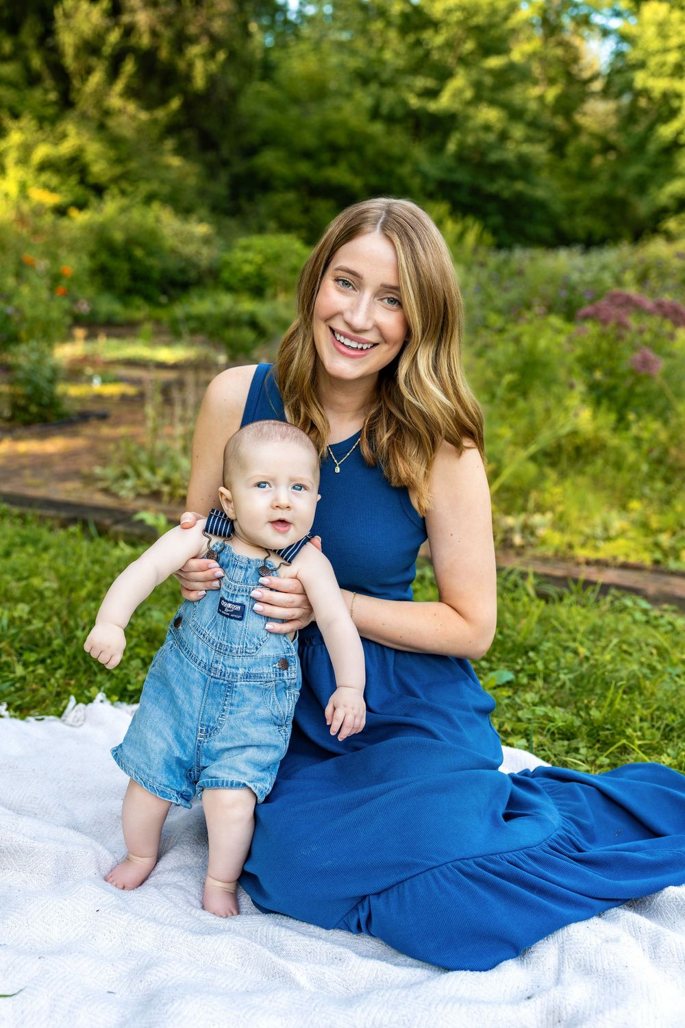 A smiling woman in a blue dress sits on a white blanket outdoors, holding a baby in denim overalls in a lush garden.