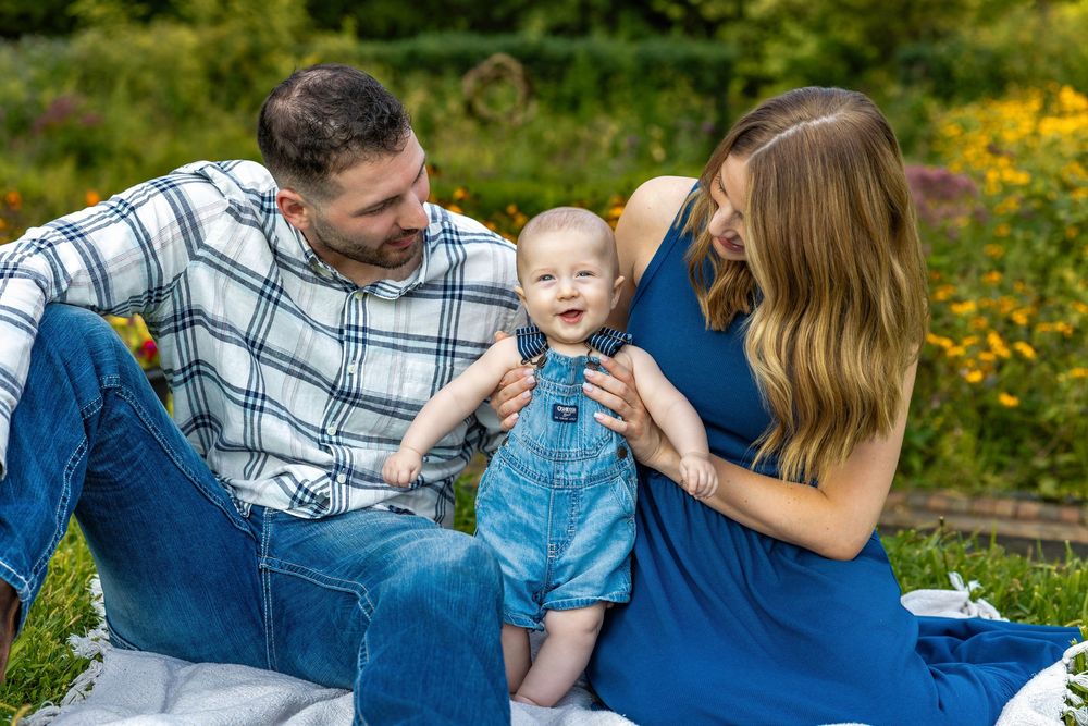 Happy family with baby in denim overalls enjoying a sunny outdoor picnic in a lush garden setting.