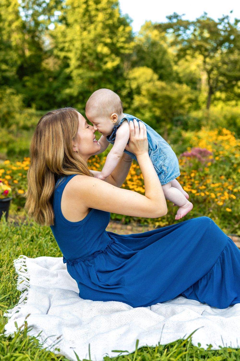 Mother in blue dress holds baby up for a nose kiss on a white blanket in a lush garden.