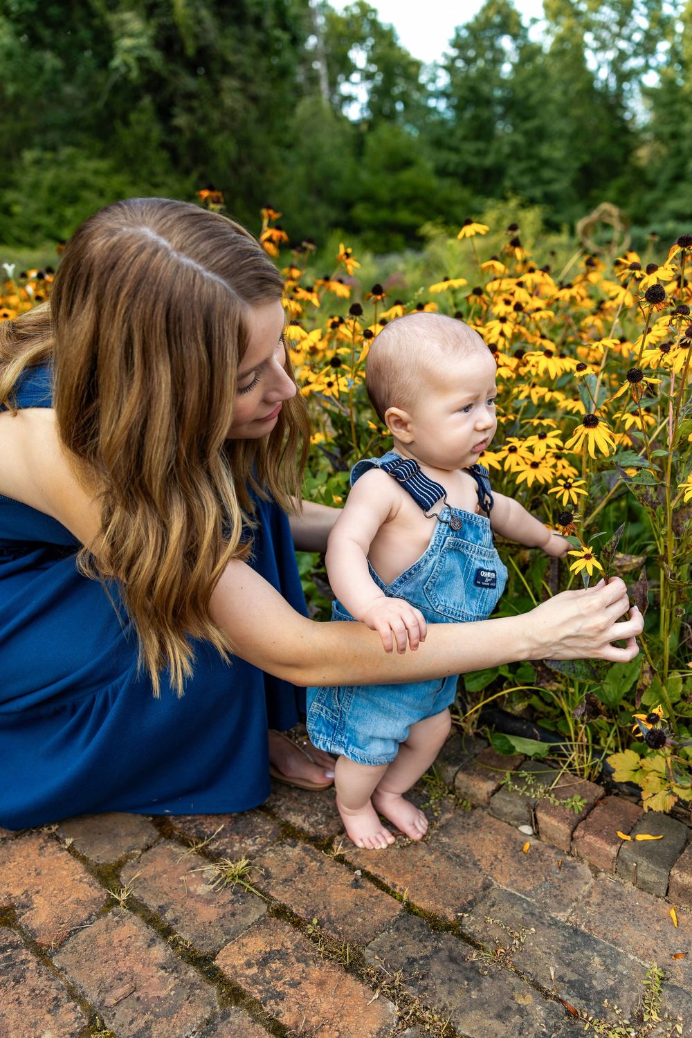 Mother in blue dress helps baby in denim overalls touch yellow black-eyed Susan flowers in a garden.