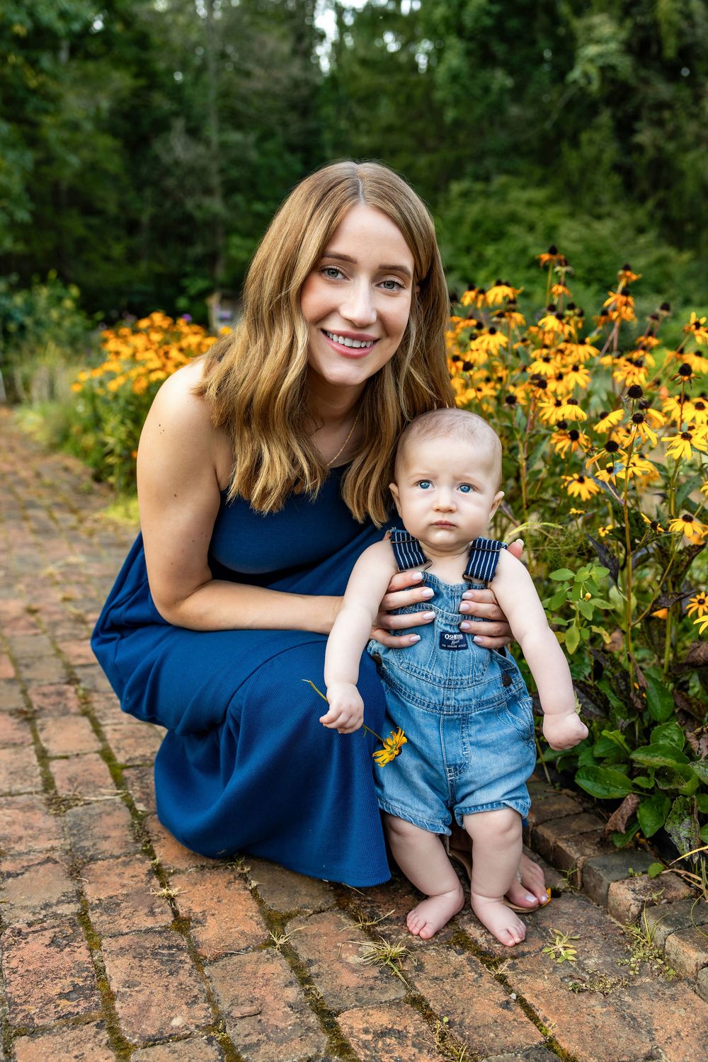 Smiling mother in blue dress crouches with baby in denim overalls by yellow flowers on brick path.
