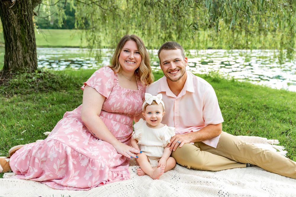 Family of three sitting on blanket by pond, smiling couple with baby girl in white outfit, lush green trees background.
