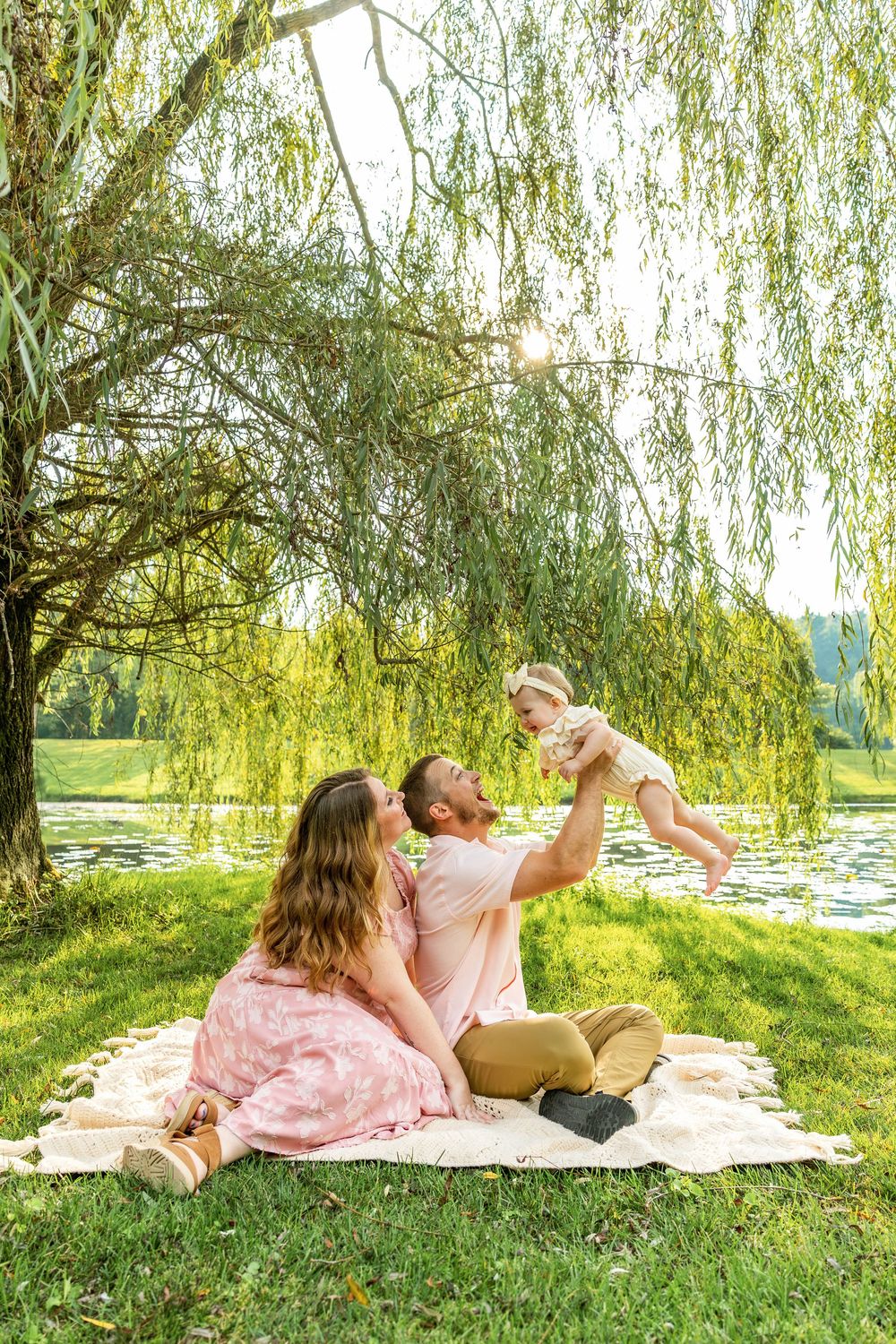 Young family sitting on blanket under willow tree by lake, father lifting baby while mother watches lovingly.
