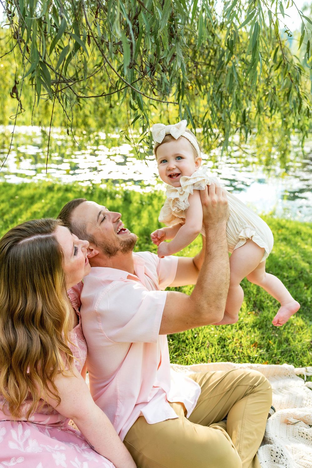 Happy family enjoying a sunny park picnic, father lifting smiling baby girl while mother looks on near a pond.