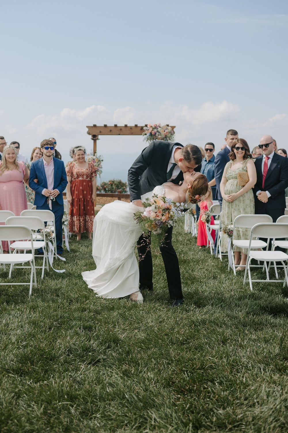 Groom dips bride for a romantic kiss in the grass aisle during an outdoor wedding ceremony.