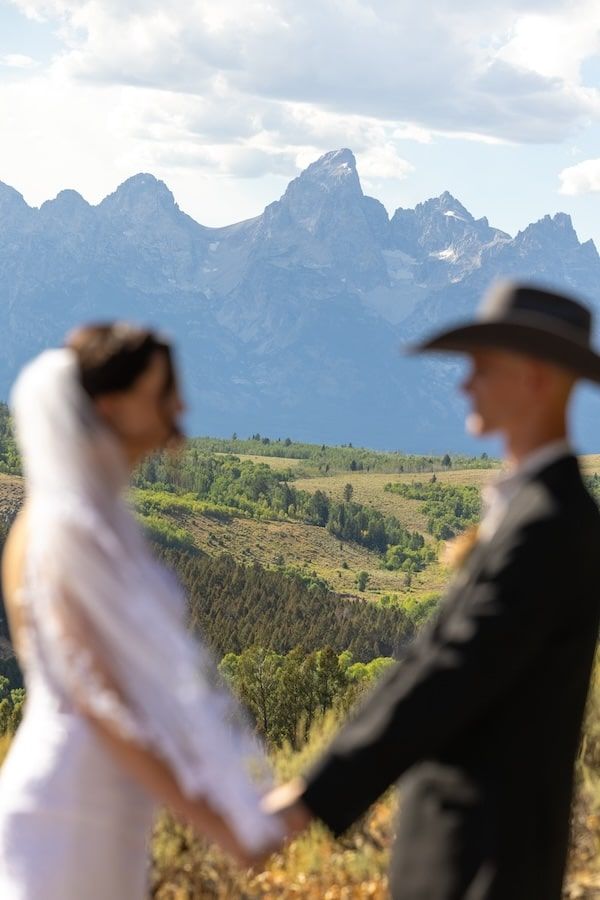 A couple poses together on a mountain ridge with dramatic Grand Teton peaks rising in the background.