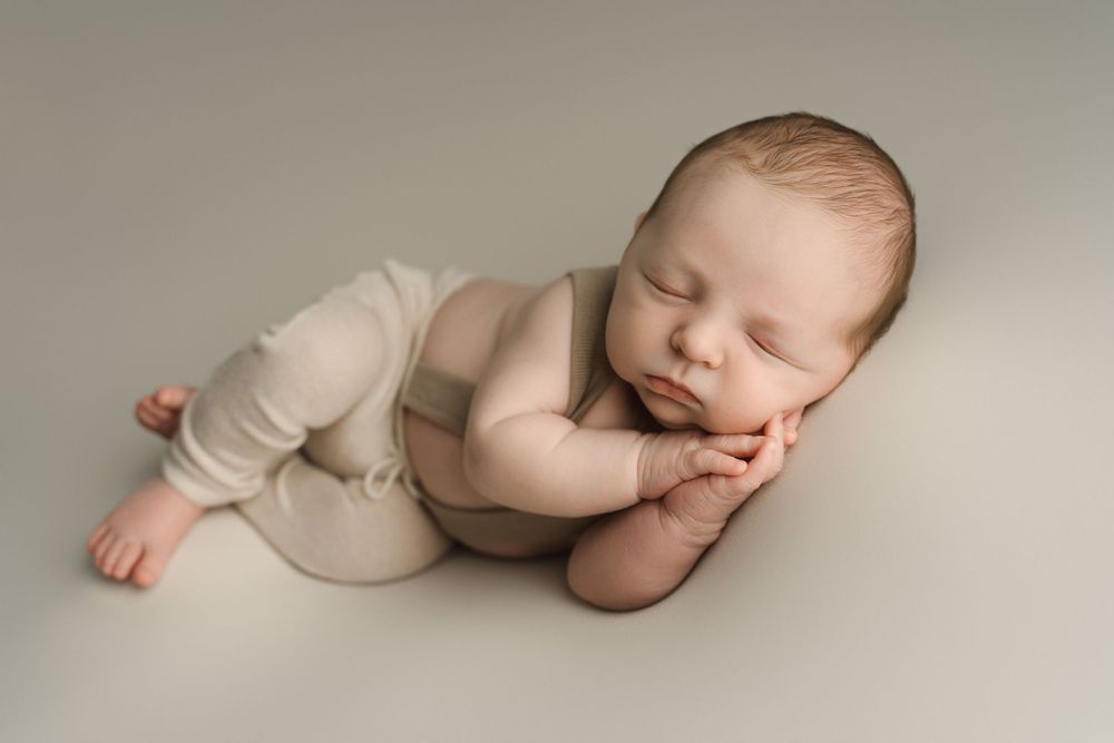 Sleeping newborn baby boy in beige romper posed with hands under chin on neutral gray backdrop.