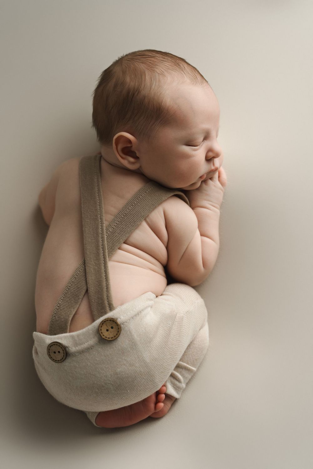 Newborn baby boy sleeping in cream suspender shorts and pink onesie on soft white background.