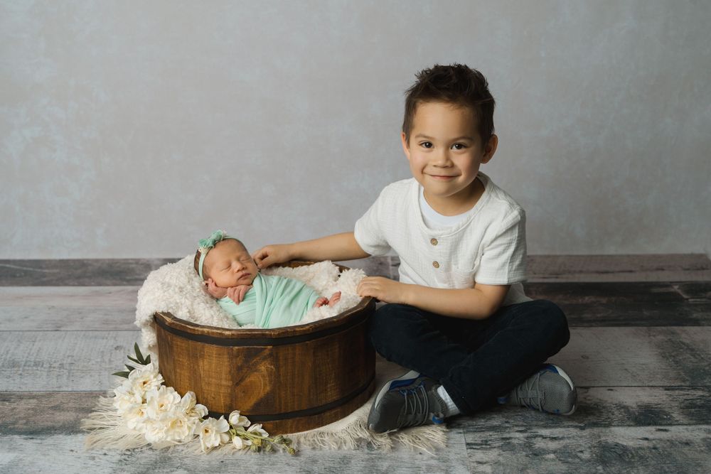 Smiling young boy sitting beside wooden basket holding swaddled newborn baby sister with white flowers.