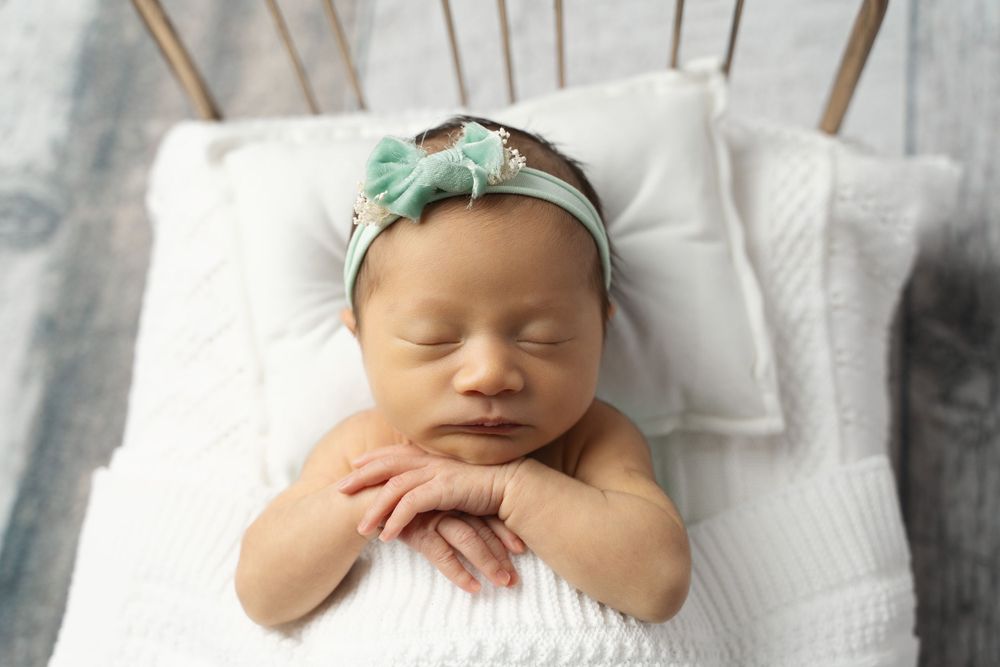 Sleeping newborn baby girl with mint green bow headband resting hands on white knit blanket.