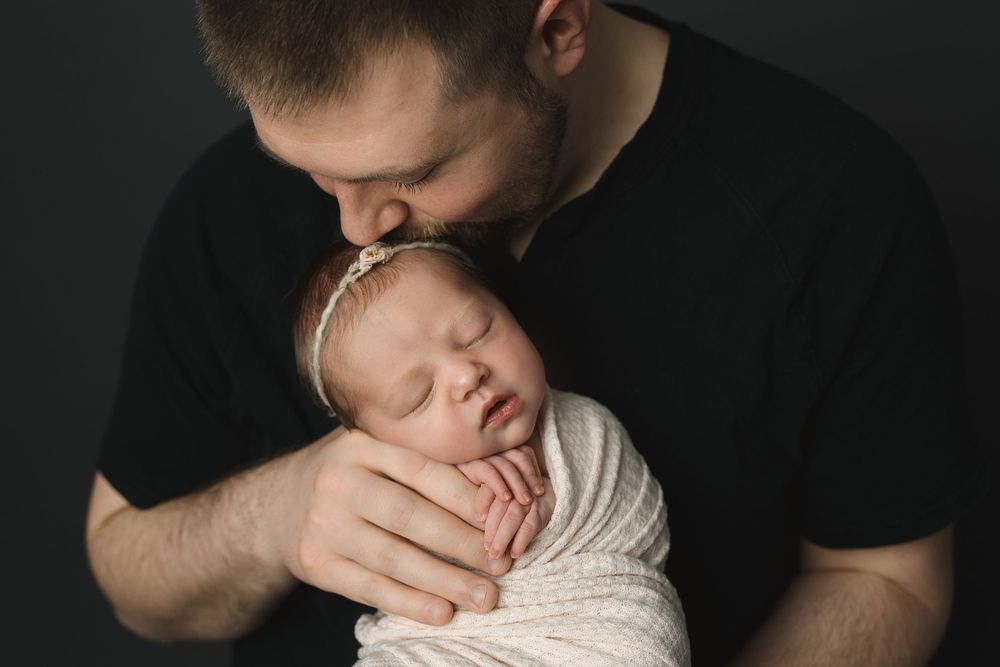 Loving father in a black shirt gently kissing his sleeping swaddled newborn baby.