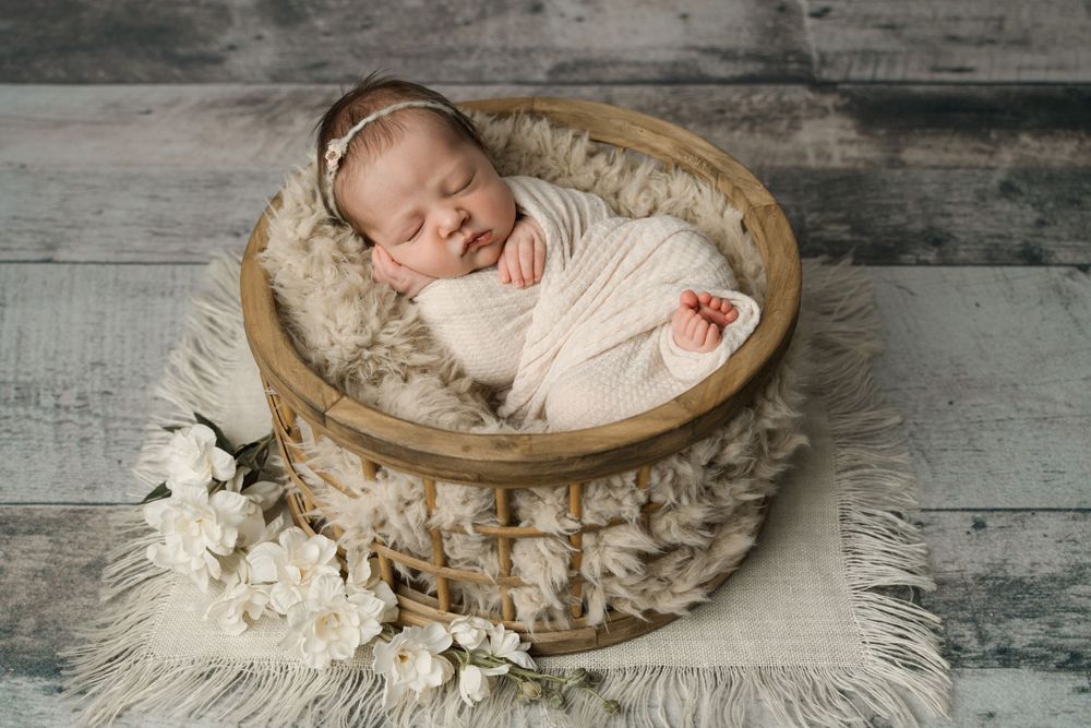 Peaceful newborn baby girl swaddled in cream wrap resting in wooden basket with fur and white flowers.