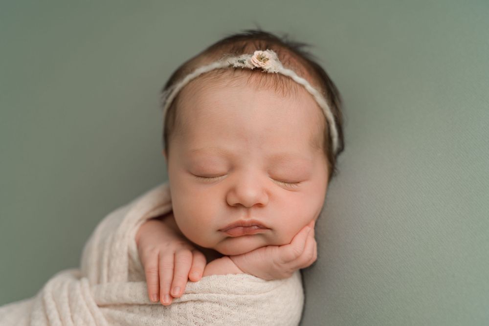 Sleeping newborn baby girl wrapped in white knit swaddle with floral headband on sage green background.